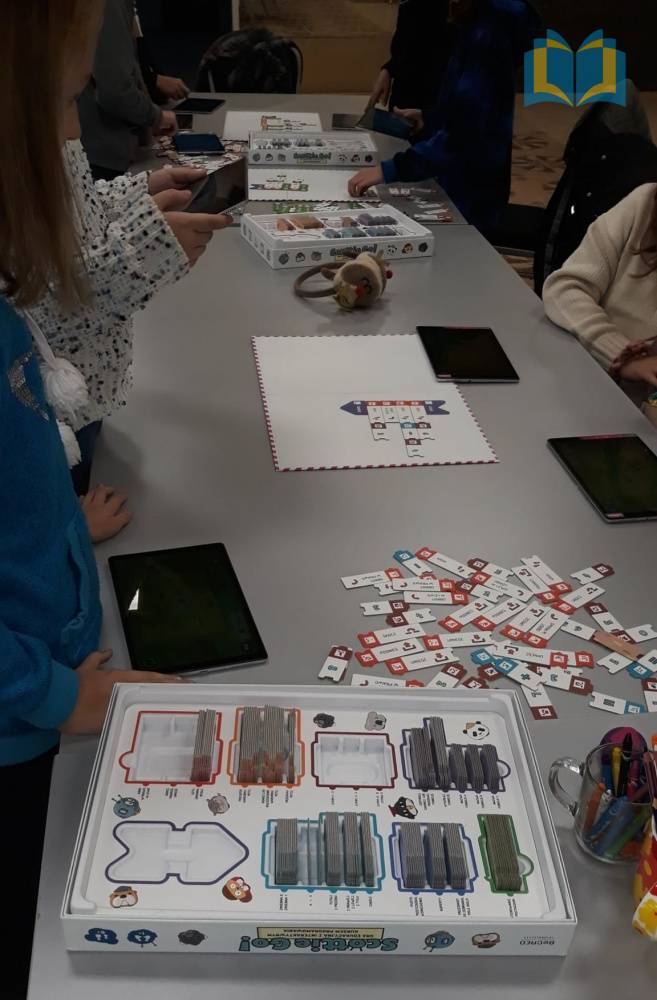 Zdjęcie: Dzieci uczestniczą w zajęciach z programowania. Stoją przy stole, na którym znajduje się gra edukacyjna Scottie Go. Za pomocą tabletów uczą się programowania.